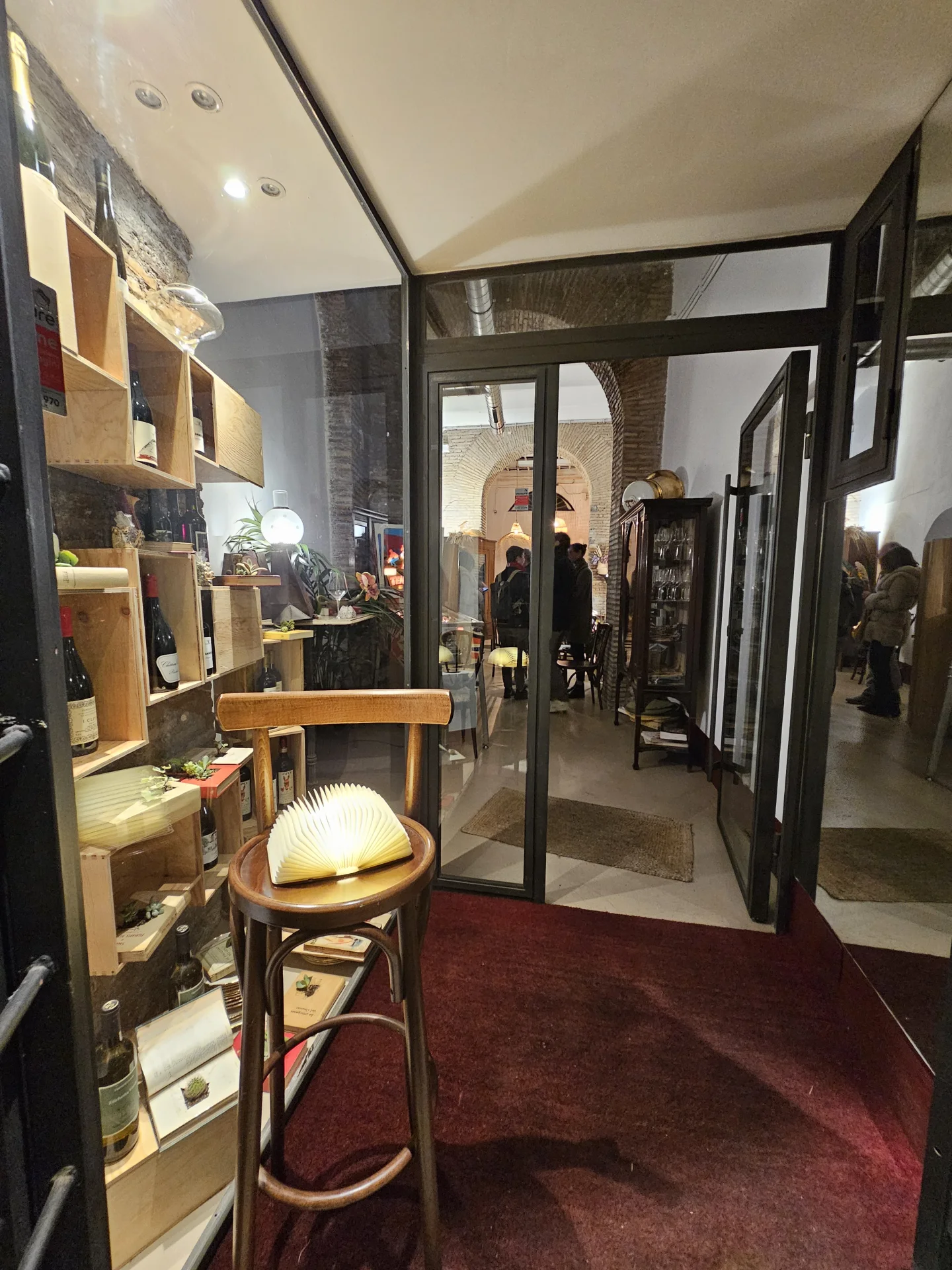 Eingang von [[nt]]Accanto [[/nt]] mit Regalen mit Naturweinen, Bücherlampe und Blick auf den Backsteinbogen – Caffè Novecento Roma